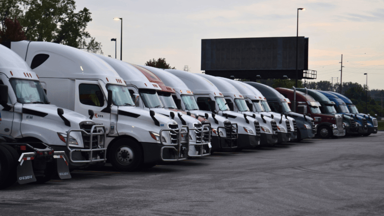 A long row of semitruck cabs, most of them white with a few red, green and blue, parked nose-out in a truck stop lot at dawn, with trees, light poles and a billboard behind them.