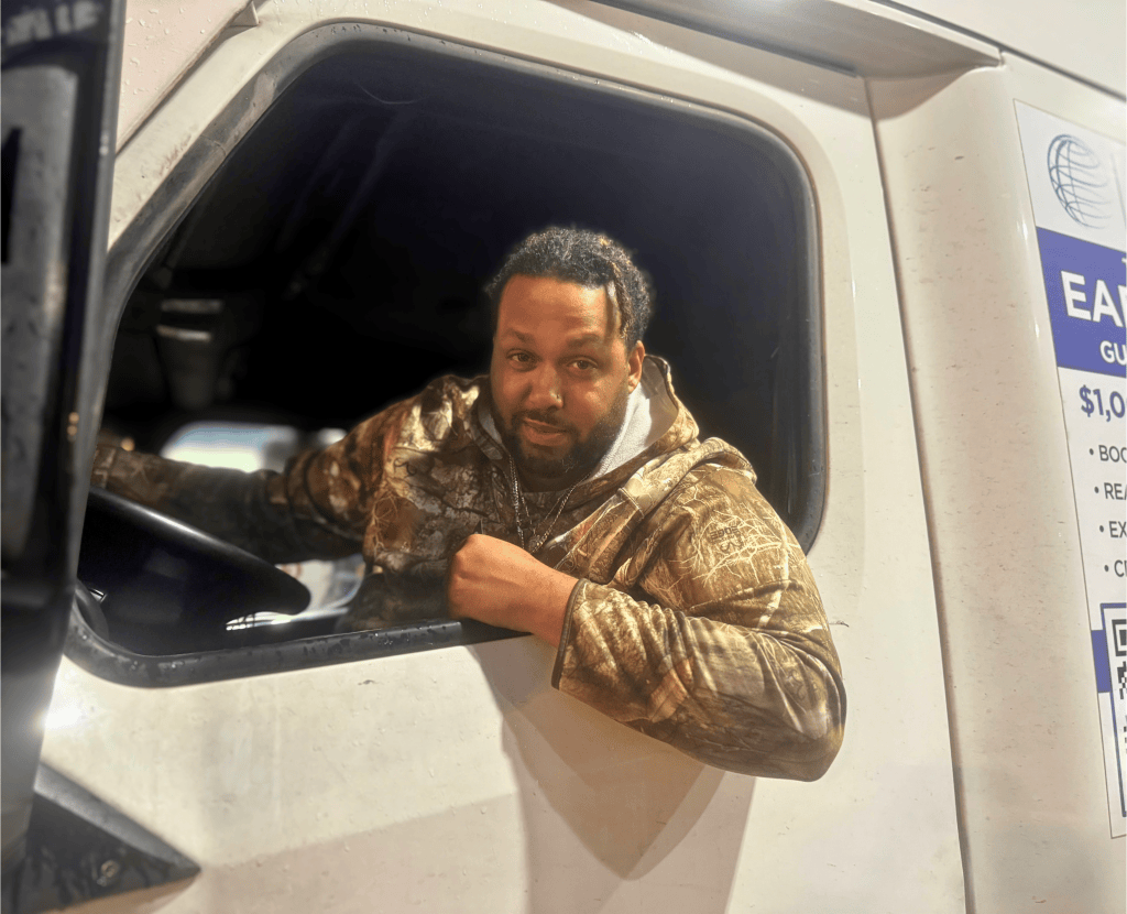 A man with curly hair and a beard, wearing a camouflage hoodie, leans out the driver's-side window of a white semitruck at night, one hand on the steering wheel.