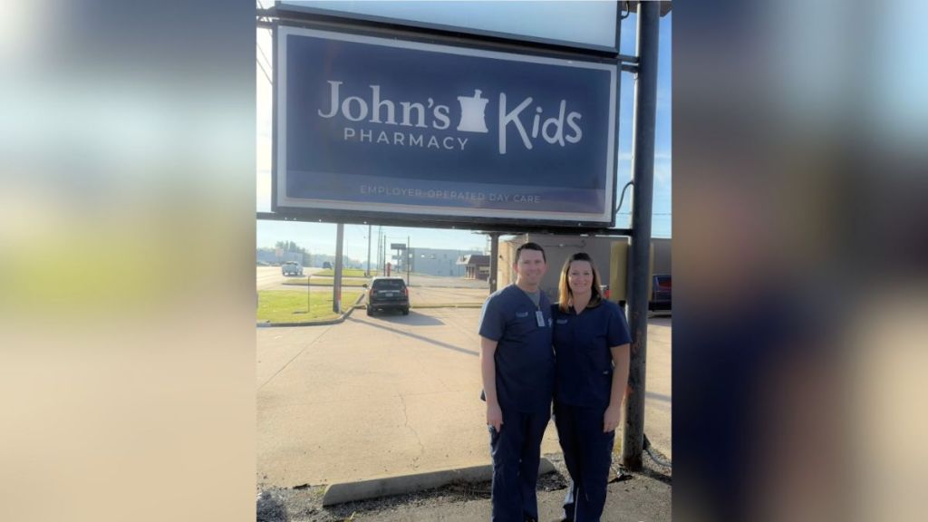 Abe Funk, the owner of John's Pharmacy, and his wife, stand in front of a sign for the pharmacy's daycare. Missouri is investing nearly $1.5 million to expand a childcare cost-sharing program to rural health employers.